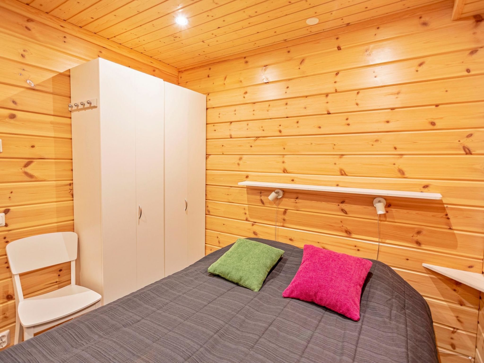 Een kamer met houten wanden en een bed met twee kussens. Er is een witte kledingkast en een stoel.