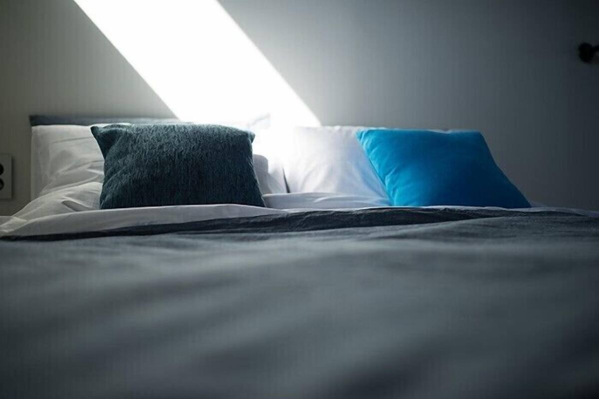 A neatly made bed with a dark blue pillow and a white pillow, a black bedspread, and a white wall with a window casting light on the bed.