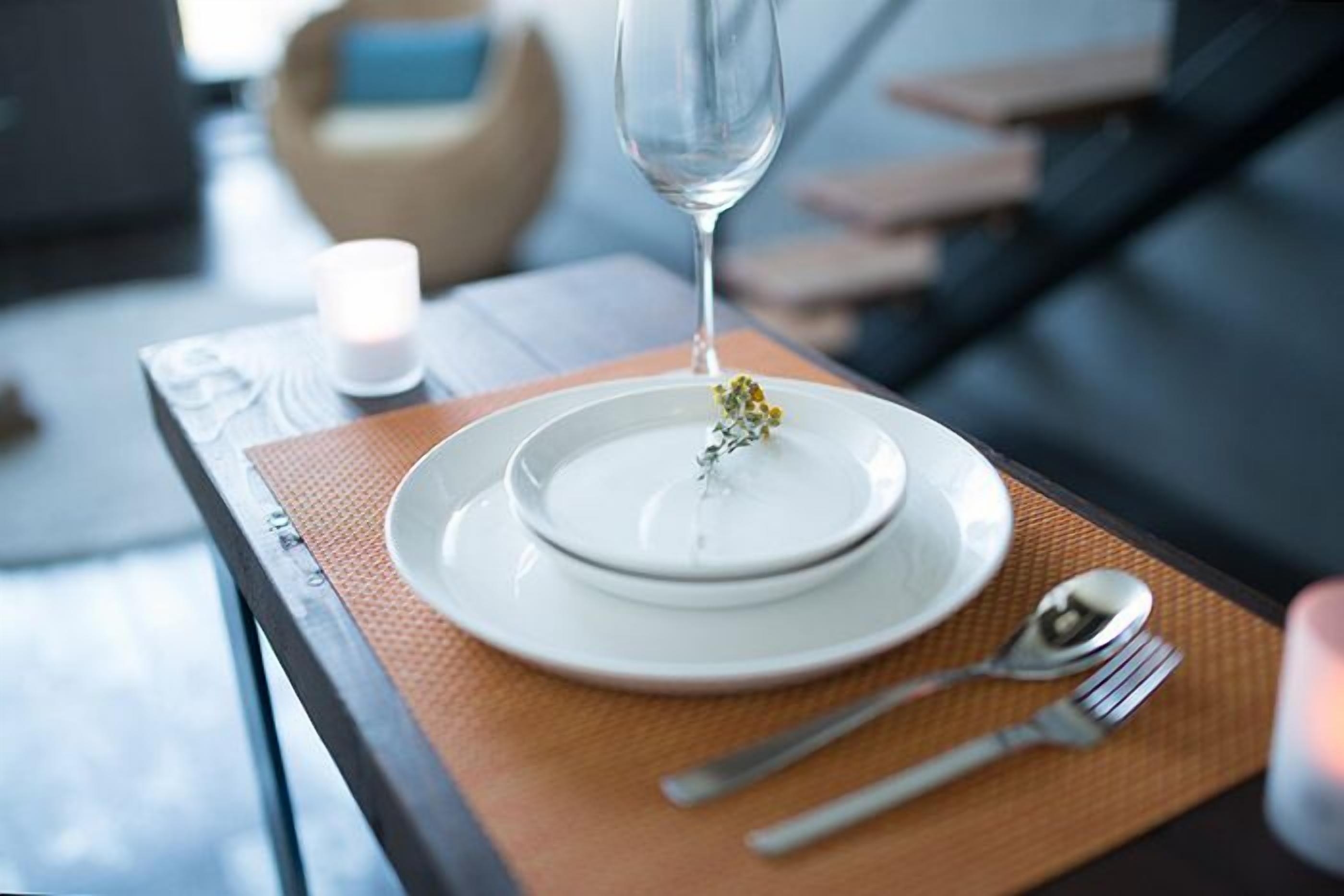 Una mesa con un plato blanco, un vaso de agua y una cuchara sobre un posavasos tejido.