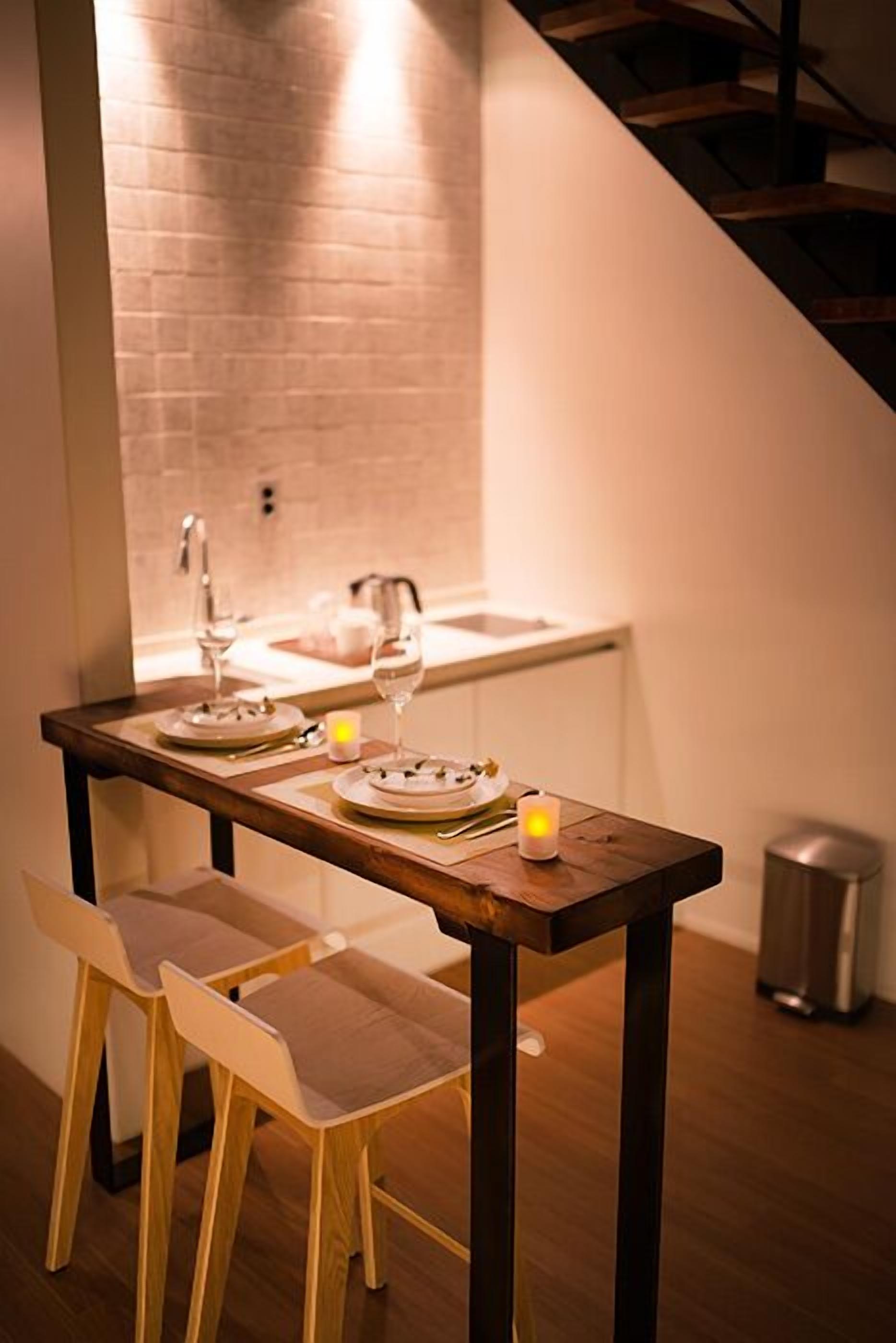 A dining area with a wooden table set for two, a small candle, and a view of a kitchen in the background.
