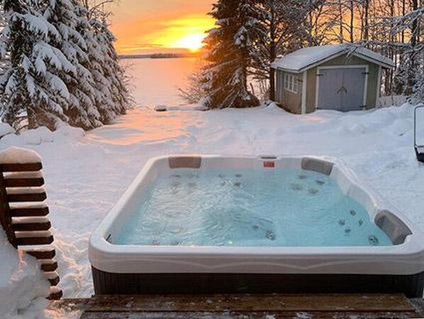 A hot tub surrounded by snow-covered trees and a small shed, with a sunset in the background.