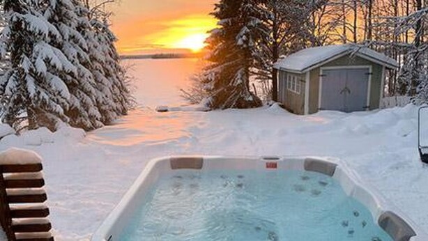 Ein Whirlpool, umgeben von schneebedeckten Bäumen und einer kleinen Werkstatt, mit einem Sonnenuntergang im Hintergrund.
