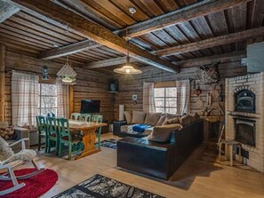 Interior rústico de cabaña con techo de madera, chimenea, sofá, mesa de comedor con sillas y televisor.