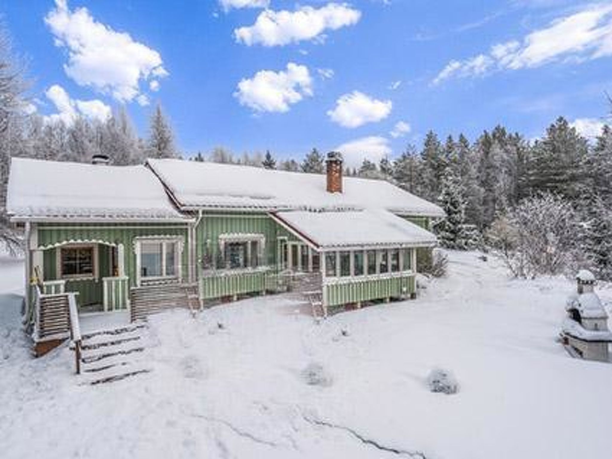 A green wooden house with a snow-covered roof and porch, surrounded by trees.