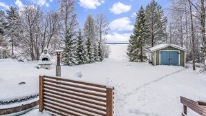 Eine verschneite Landschaft mit einer hölzernen Bank, einem kleinen Schuppen und schneebedeckten Bäumen.
