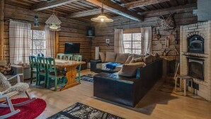Ein rustikales Blockhaus-Interieur mit Holzbalkendecke, Kamin, einer Couch, einem Esstisch mit Stühlen und einem Fernseher.