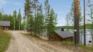 Une cabane située au bout d’un chemin de terre, entourée de grands arbres et bordée par un lac.