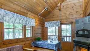 Un intérieur de chalet chaleureux avec des murs en bois, une cheminée en pierre, un coin repas avec une table et des bancs, et de grandes fenêtres offrant une vue sur l’extérieur.