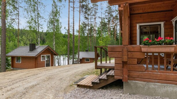 Une cabane en bois dotée d’un porche et d’une petite terrasse, entourée d’une allée en gravier et d’une forêt dense.