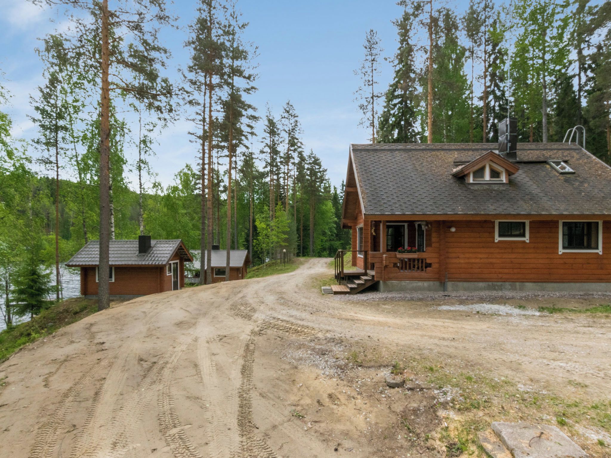 Eine Holzhütte mit einer unbefestigten Auffahrt, die zu einer Holzterrasse führt, umgeben von hohen Bäumen und einem Gewässer.