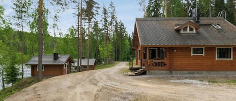 A cabin with a dirt driveway leading to a wooden deck, surrounded by tall trees and a body of water.
