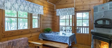 Interior de una cabaña con techo de madera, chimenea, mesa de comedor con sillas, sofá y ventanas amplias.