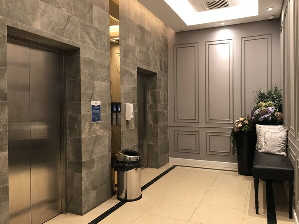 Un espace moderne avec ascenseur, doté de murs en pierre, d’une poubelle et d’un banc agrémenté d’un arrangement floral.