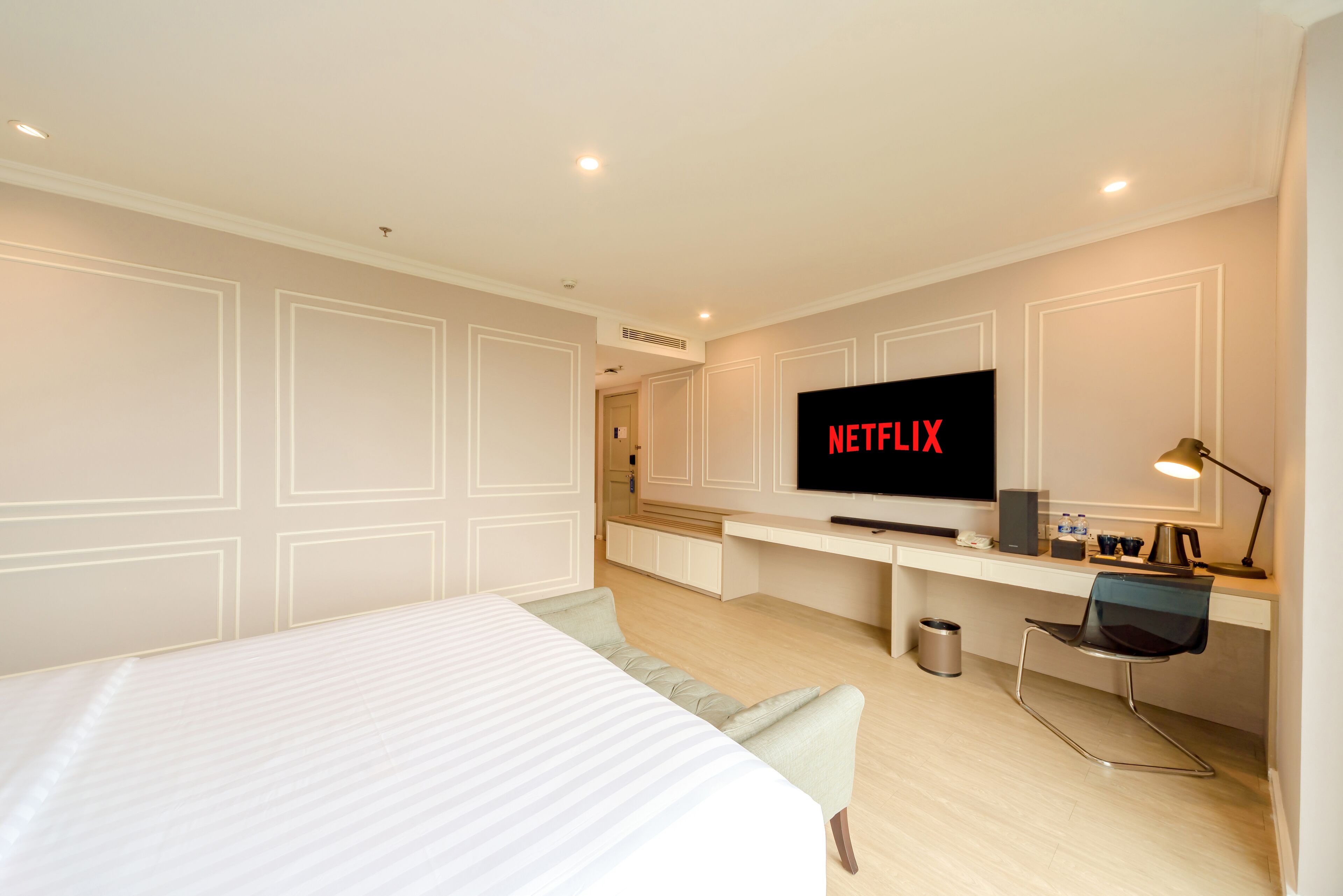 Una habitación de hotel moderna con una cama grande, un escritorio con una computadora, un televisor mostrando 'NETFLIX' y un pequeño basurero.
