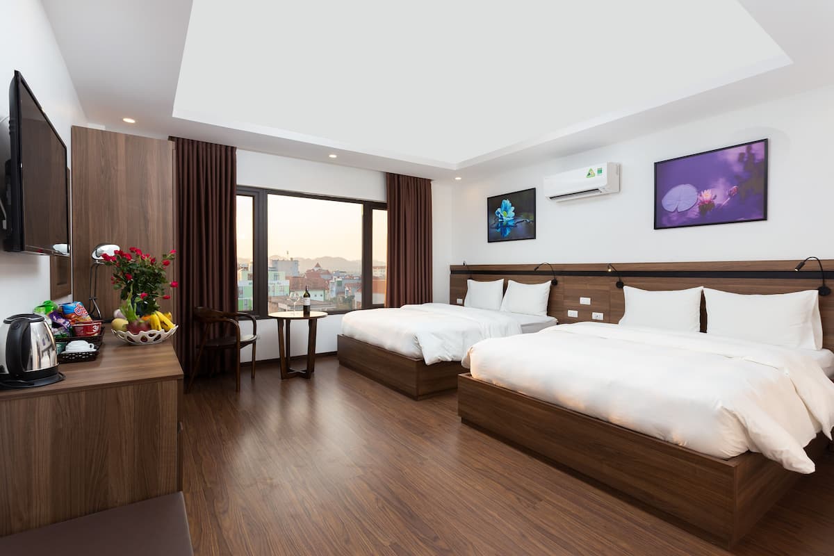 A hotel room with two beds, a desk with a kettle, a fruit bowl, and a city view.