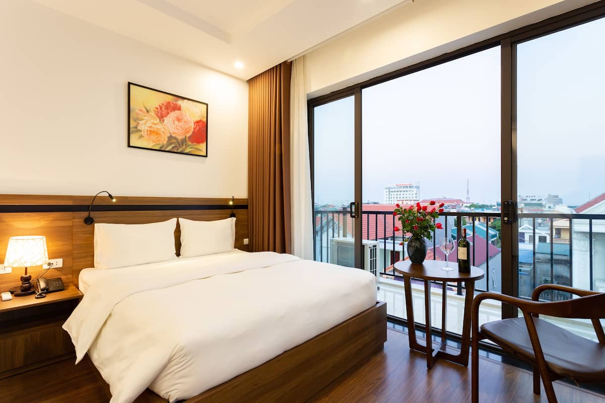 A hotel room with a large bed, a nightstand with a lamp, a chair, a table with a vase of flowers, and a balcony with a view of the city.
