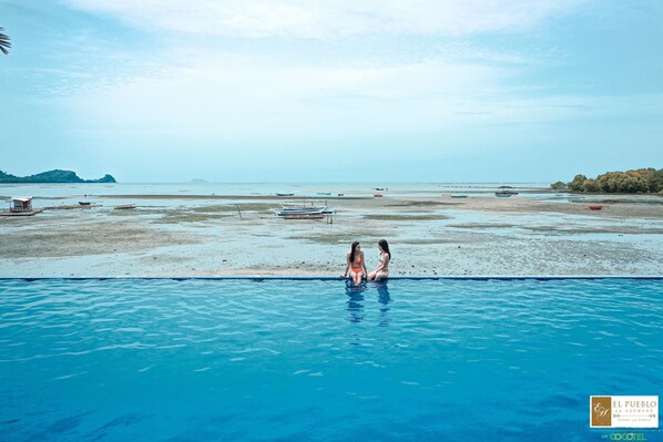 Uma piscina com duas pessoas na água, uma praia com barcos e uma ilha ao longe.