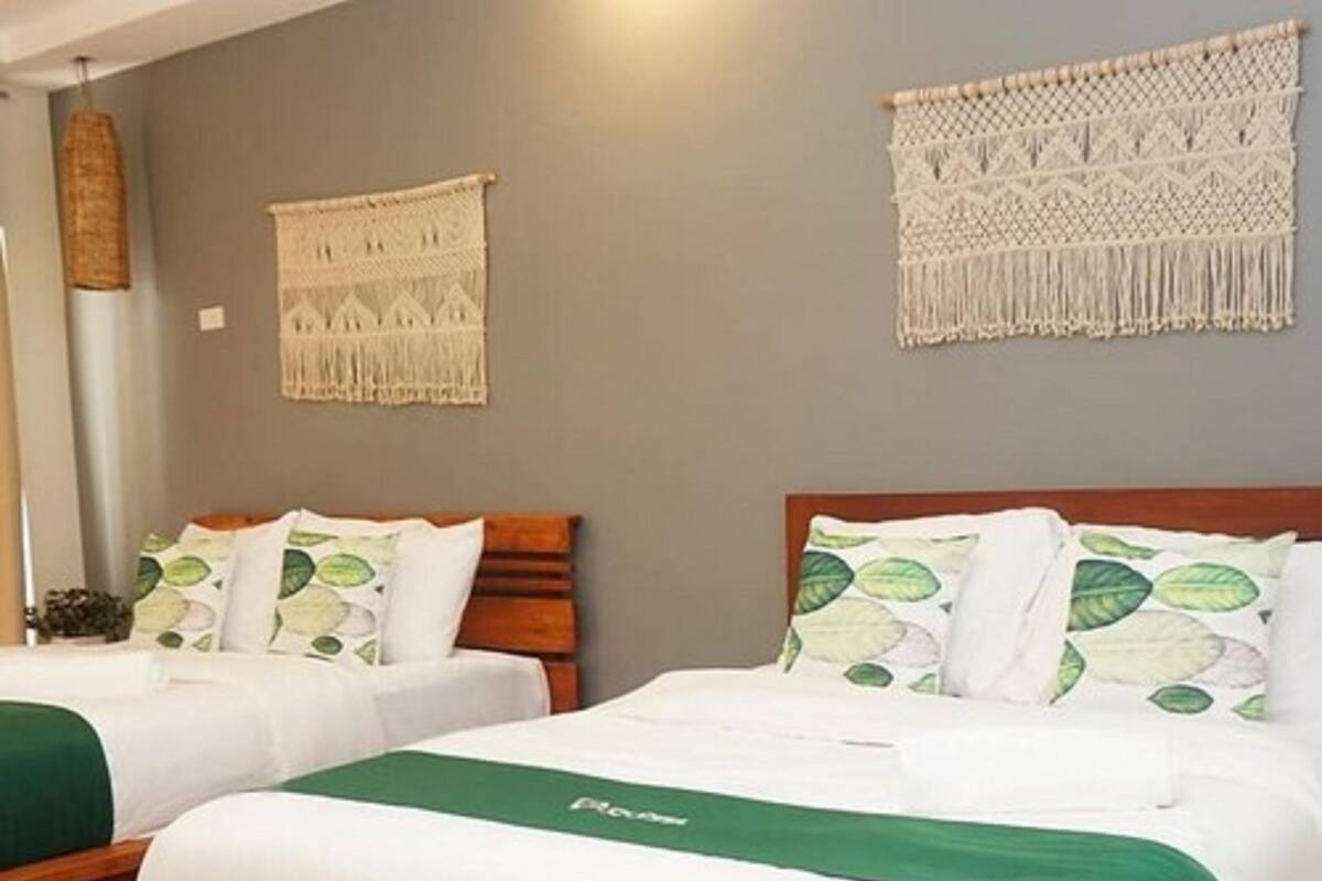 Habitación de hotel con dos camas de madera, cada una con ropa de cama blanca y almohadones verdes. Se observan tapices de macramé sobre las camas.