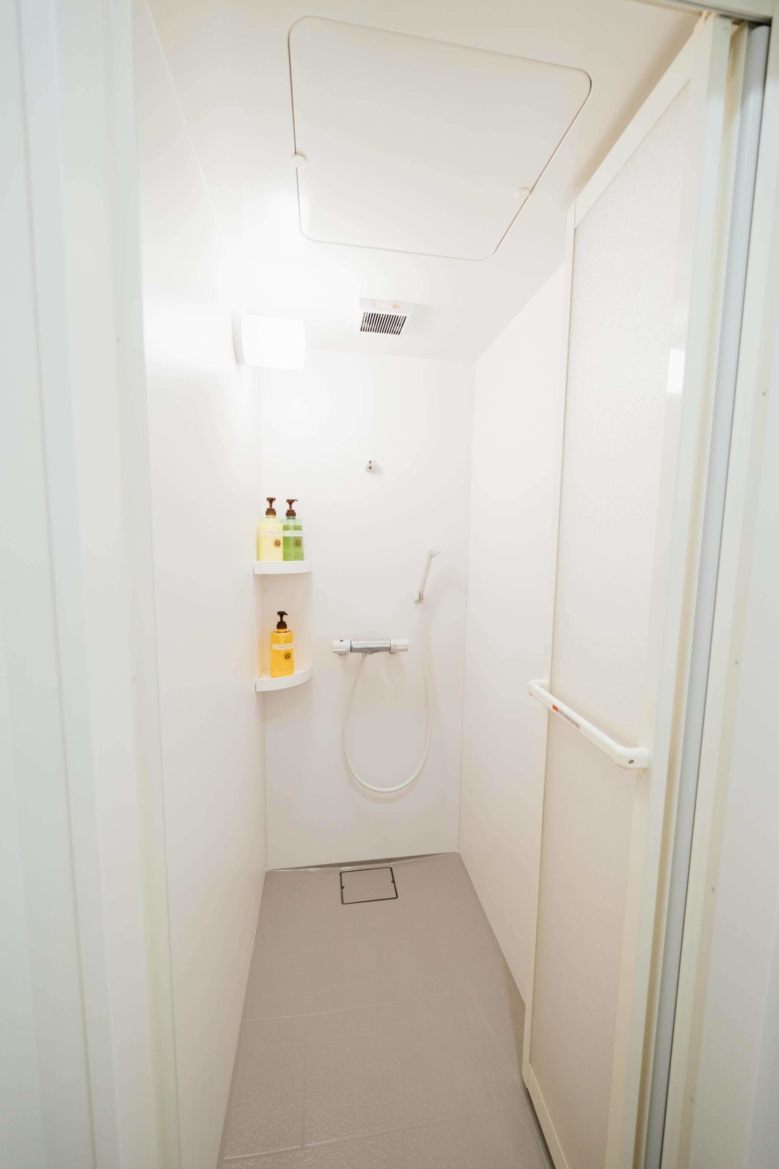 A small, white bathroom with a shower, a toilet, and a shelf with toiletries.