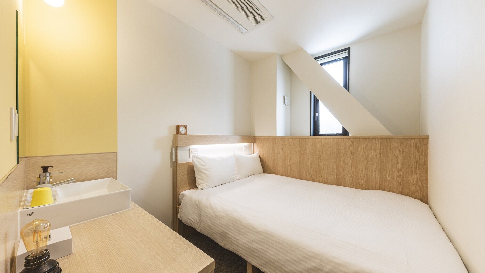 A single bed with a wooden headboard, a bedside table with a lamp, a small window, and a yellow wall.
