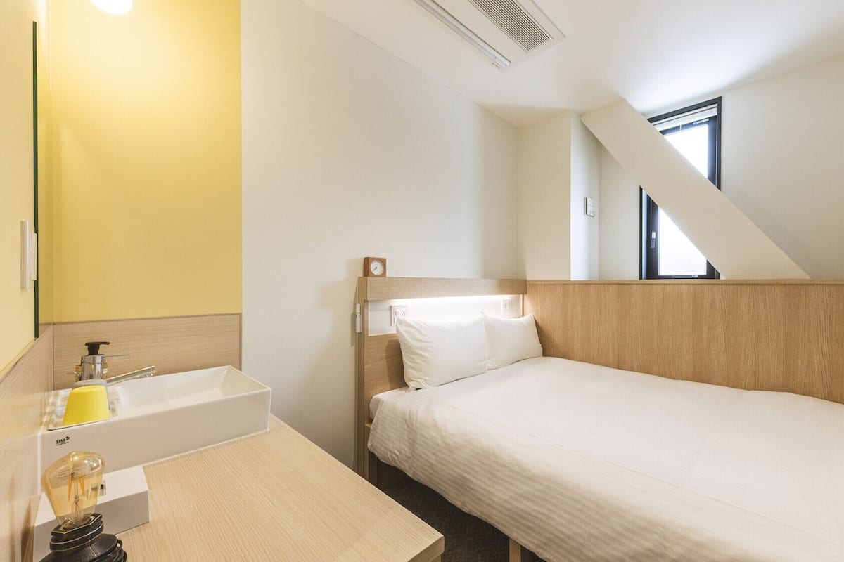 Un lit simple avec une tête de lit en bois, une table de chevet avec une lampe, une petite fenêtre et un mur jaune.