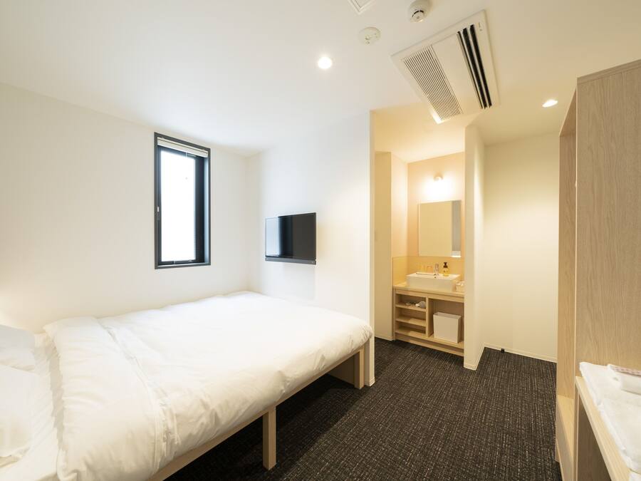 A hotel room with a single bed, a TV mounted on the wall, a window, a bathroom with a sink and mirror, and a shower area.
