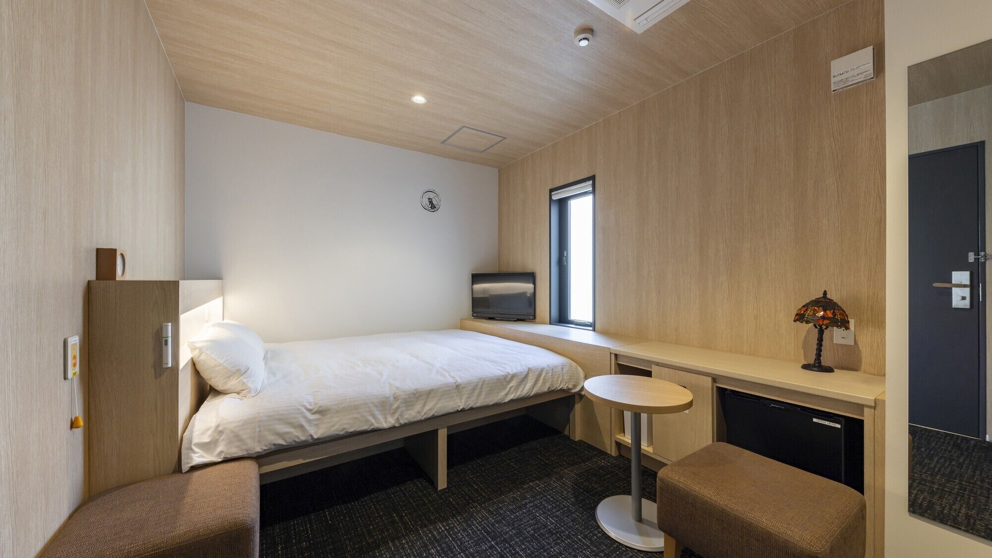 A hotel room with a bed, a desk, a chair, a small table, a lamp, a TV, and a wardrobe.