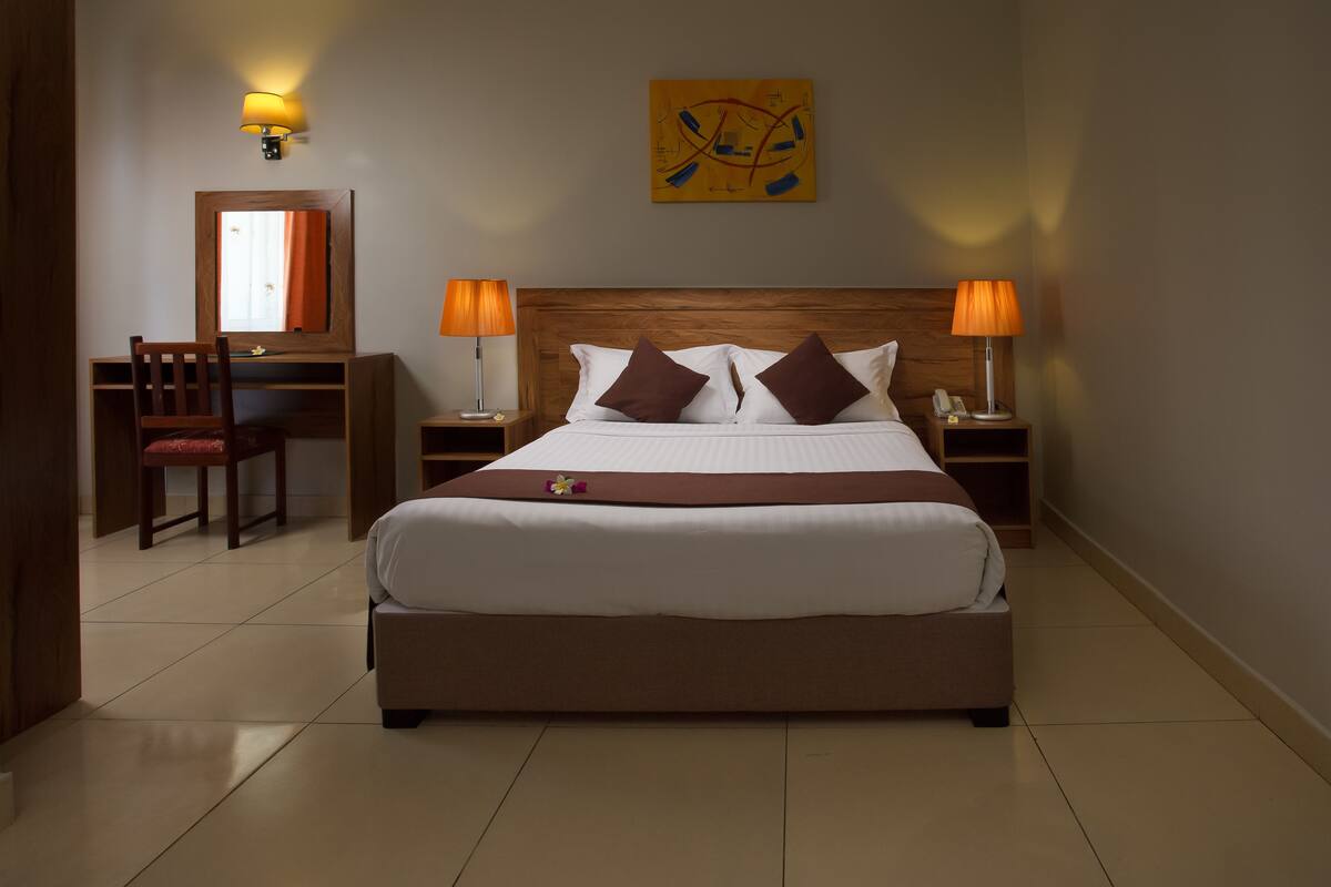 Quarto de hotel bem arrumado, com uma cama grande, duas mesas de cabeceira com abajures, uma escrivaninha com cadeira e um espelho.