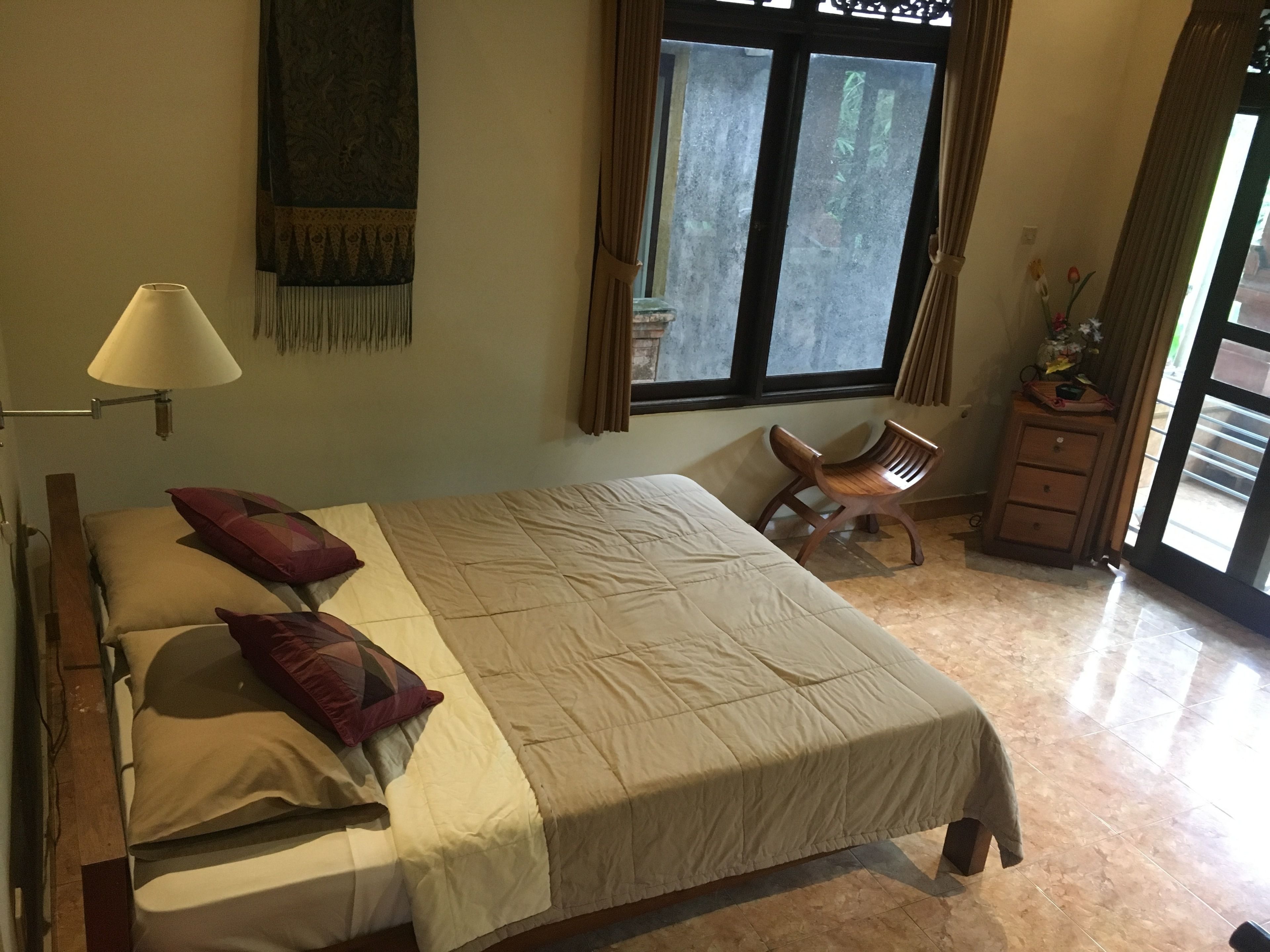 A neatly made bed with a beige bedspread and two red pillows, a wooden chair, a small wooden cabinet, and a lamp on a bedside table.