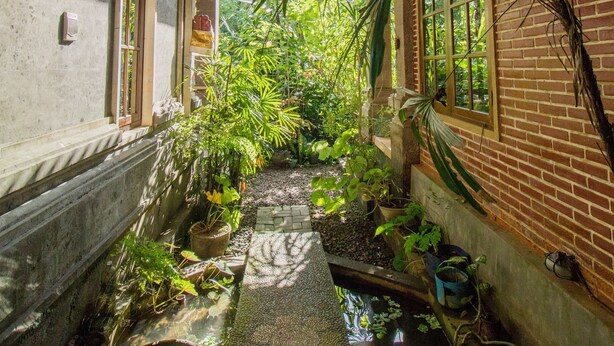 A narrow corridor with a brick wall on one side and a wooden ceiling on the other, lined with potted plants and a small water feature. - Bali Asli Lodge by EPS (Ubud)