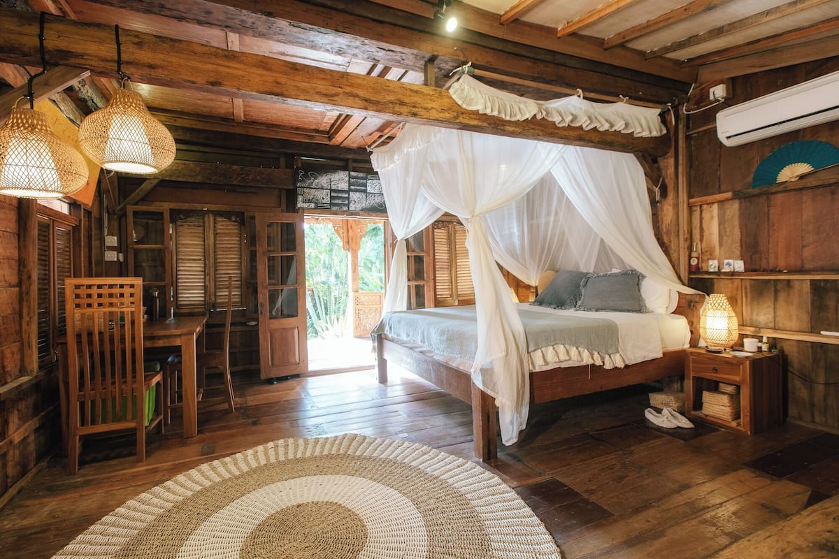 A wooden bedroom with a canopy bed, a woven rug, and a nightstand with a lamp.