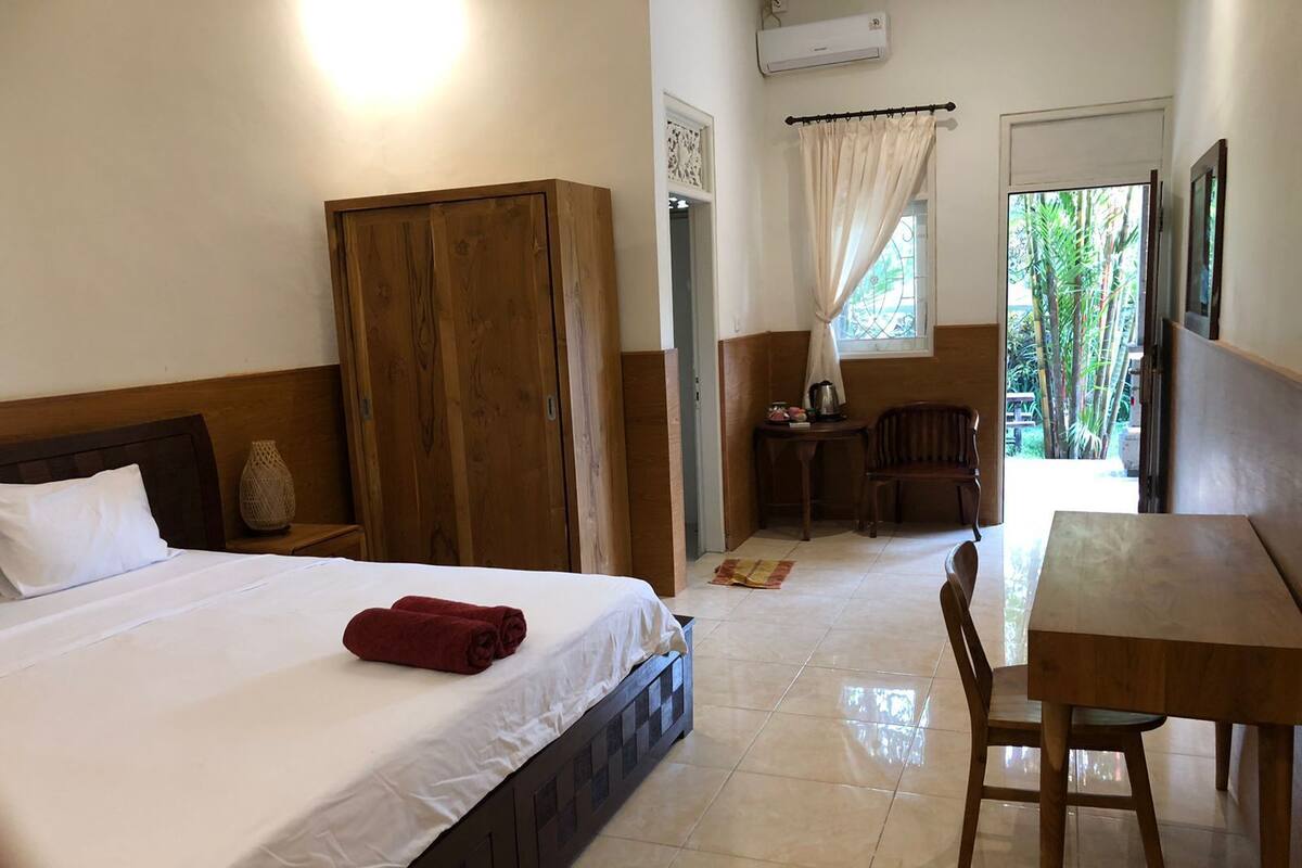 A hotel room with a bed, a wooden wardrobe, a small table with a chair, and a window with curtains.