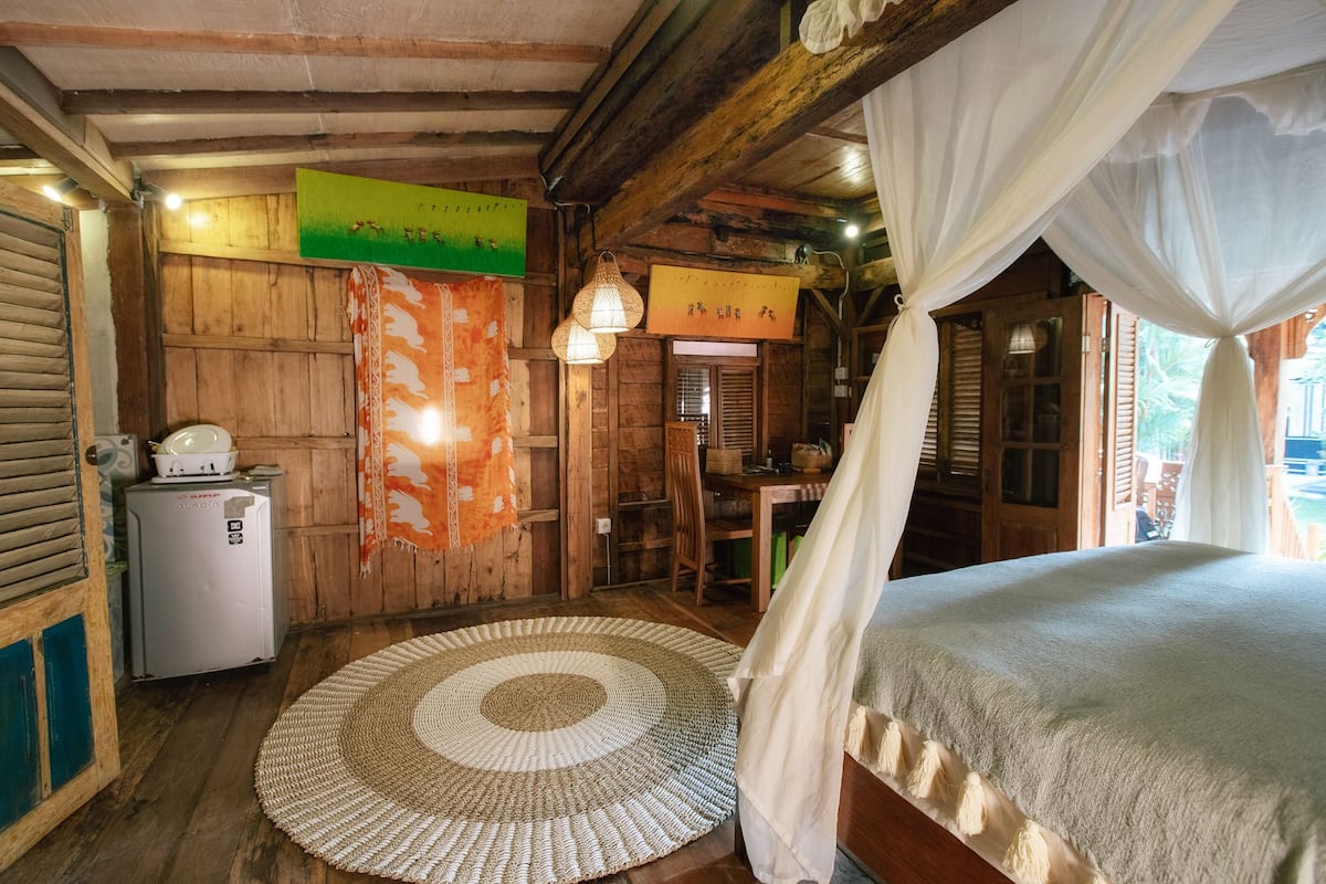 A rustic room with a bed, a small table, a chair, a woven rug, and wooden walls.