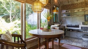 A rustic kitchen with a round wooden table, a hanging light fixture, and a view of greenery outside. - Pande Homestay (Canggu)