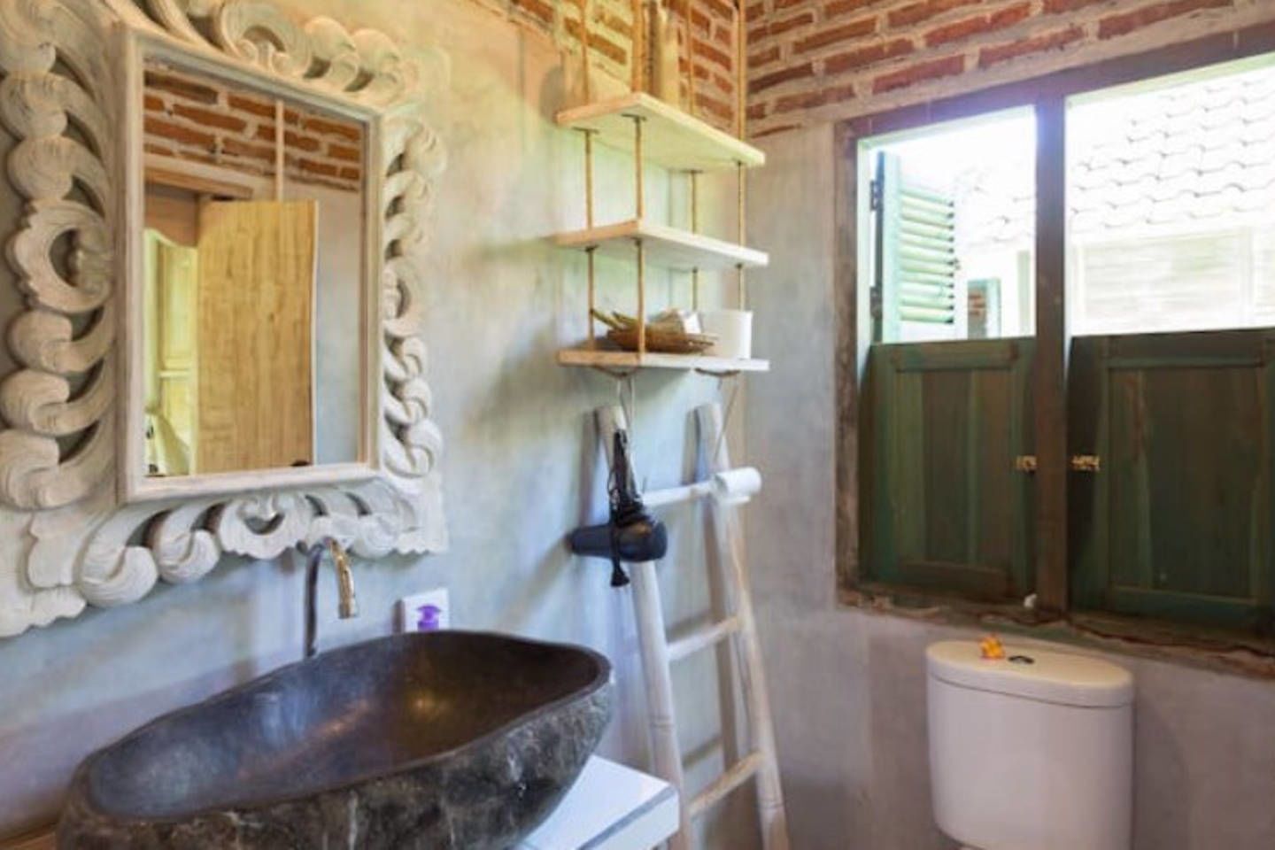 A bathroom with a large stone sink, a mirror with ornate frame, shelves with toiletries, a ladder, and a green door.