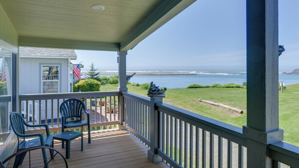 Eine Holzterrasse mit zwei Adirondack-Stühlen und einem kleinen Tisch, mit Blick auf eine Rasenfläche und das Meer.