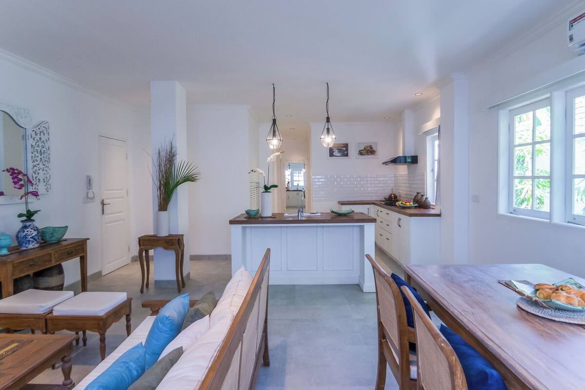Cocina moderna con armarios blancos, una isla central, iluminación colgante y un comedor con mobiliario de madera.