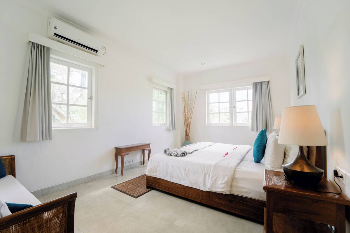 Un dormitorio con cama, mesita de noche, lámpara y ventanas.