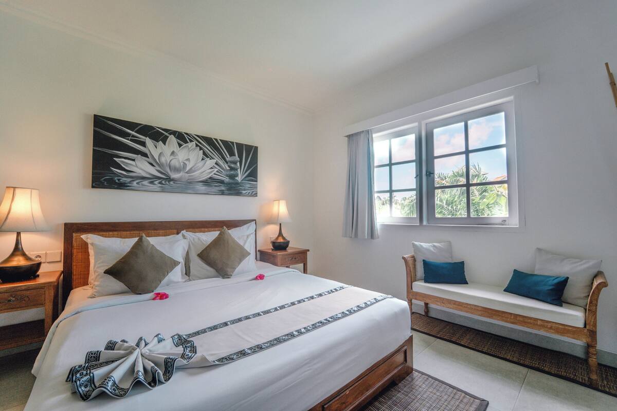Habitación de hotel ordenada con una cama grande, un banco de madera y una ventana que ofrece vistas a zonas verdes.