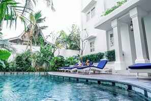 A poolside area with lounge chairs and a pool, surrounded by lush greenery and a white building with columns. - Umalas Suite (Kerobokan)
