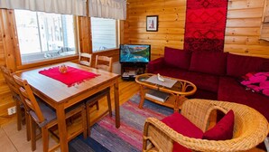 Un interior de cabaña acogedor con una mesa de comedor de madera, sillas, un televisor, un sofá rojo y una silla de mimbre.