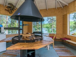 A cabin interior with a central wood-burning stove, wooden walls, and large windows offering a view of a forest and lake.