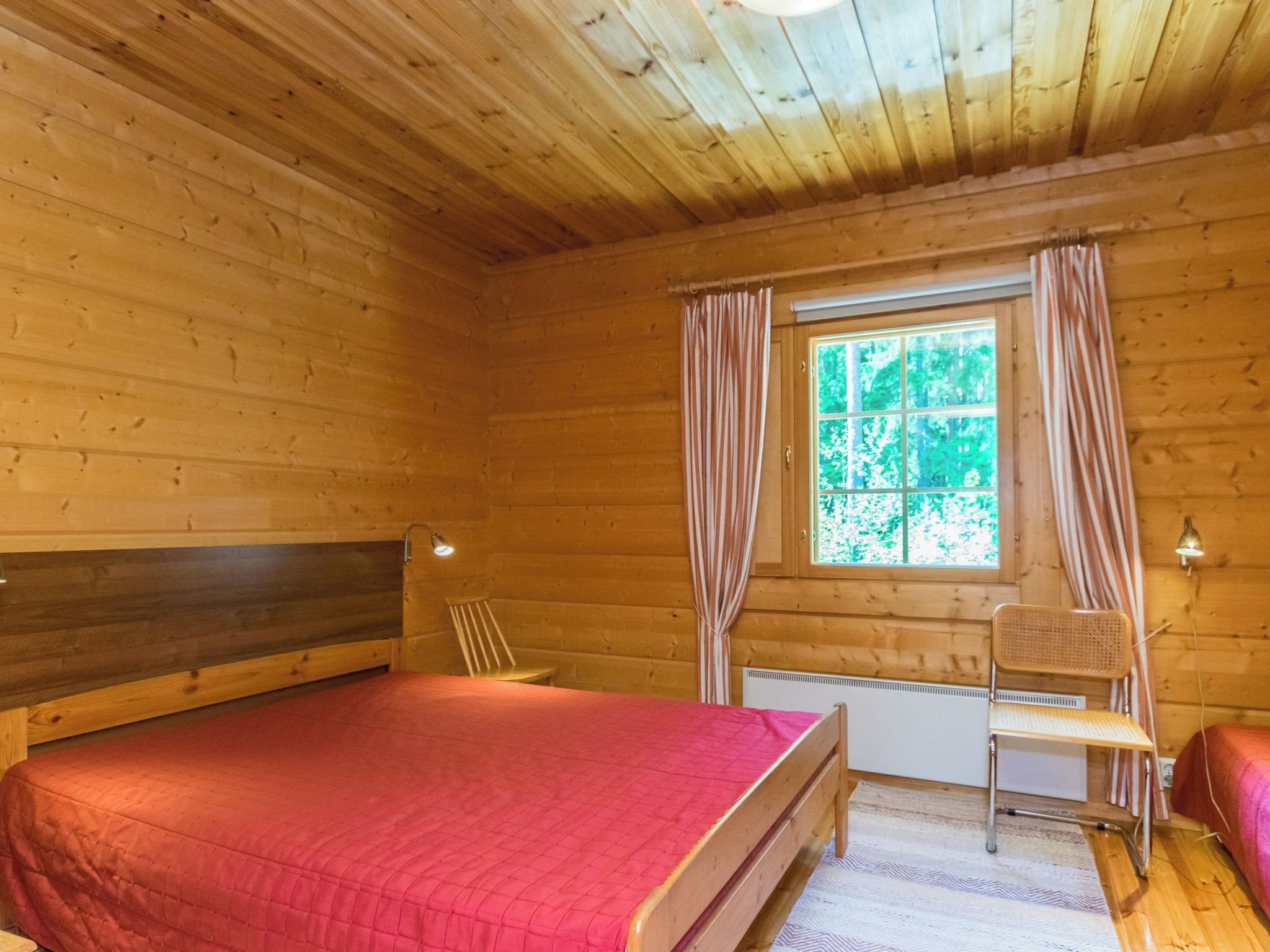 A wooden cabin room with a bed, a chair, a window with curtains, and a radiator.