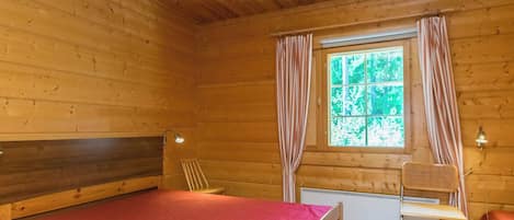A wooden cabin room with a bed, a chair, a window with curtains, and a radiator.