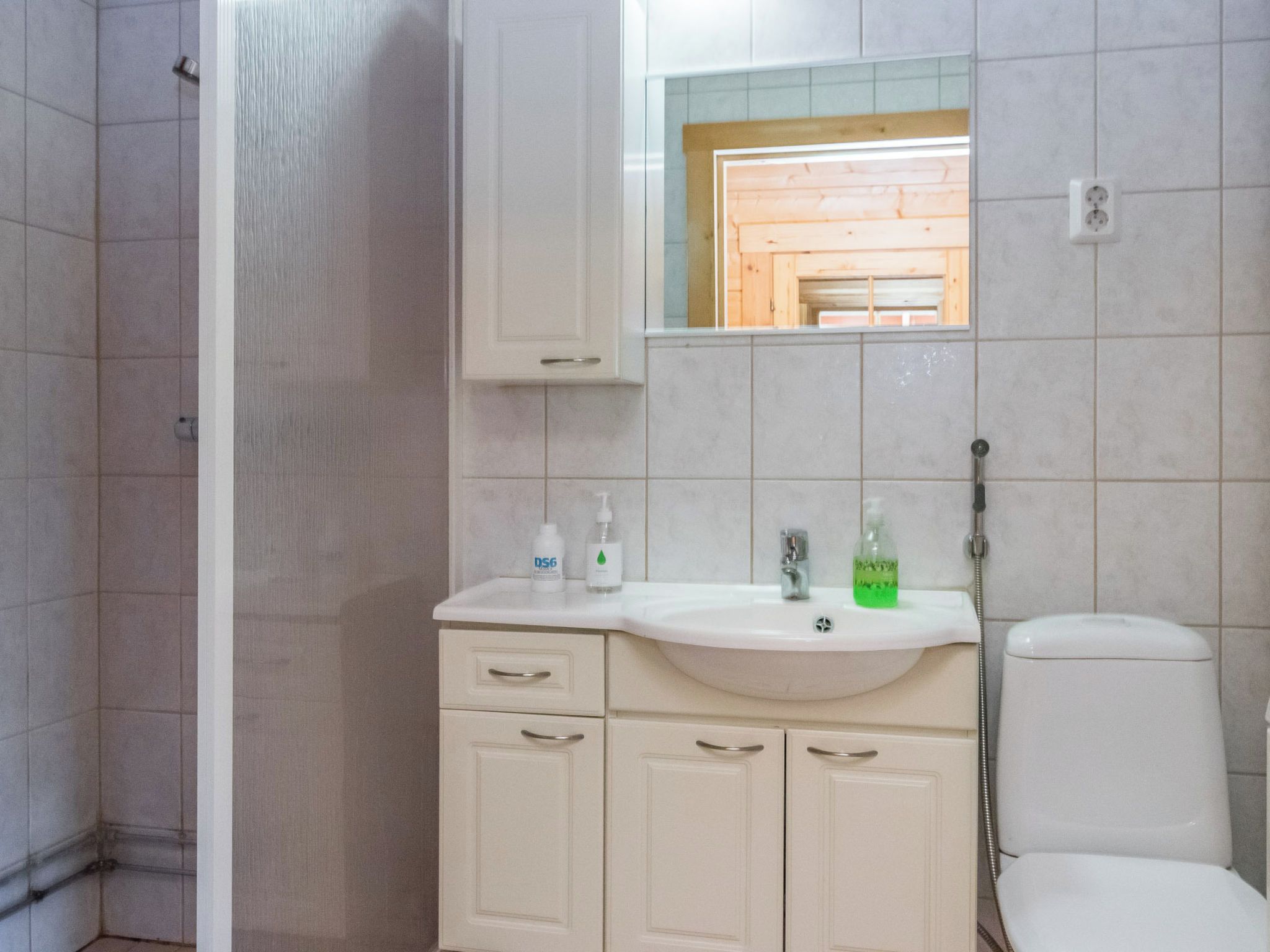 Et badeværelse med en hvid vask, spejl og toilet. Der er toiletartikler på vasken, og et bruseområde til venstre.