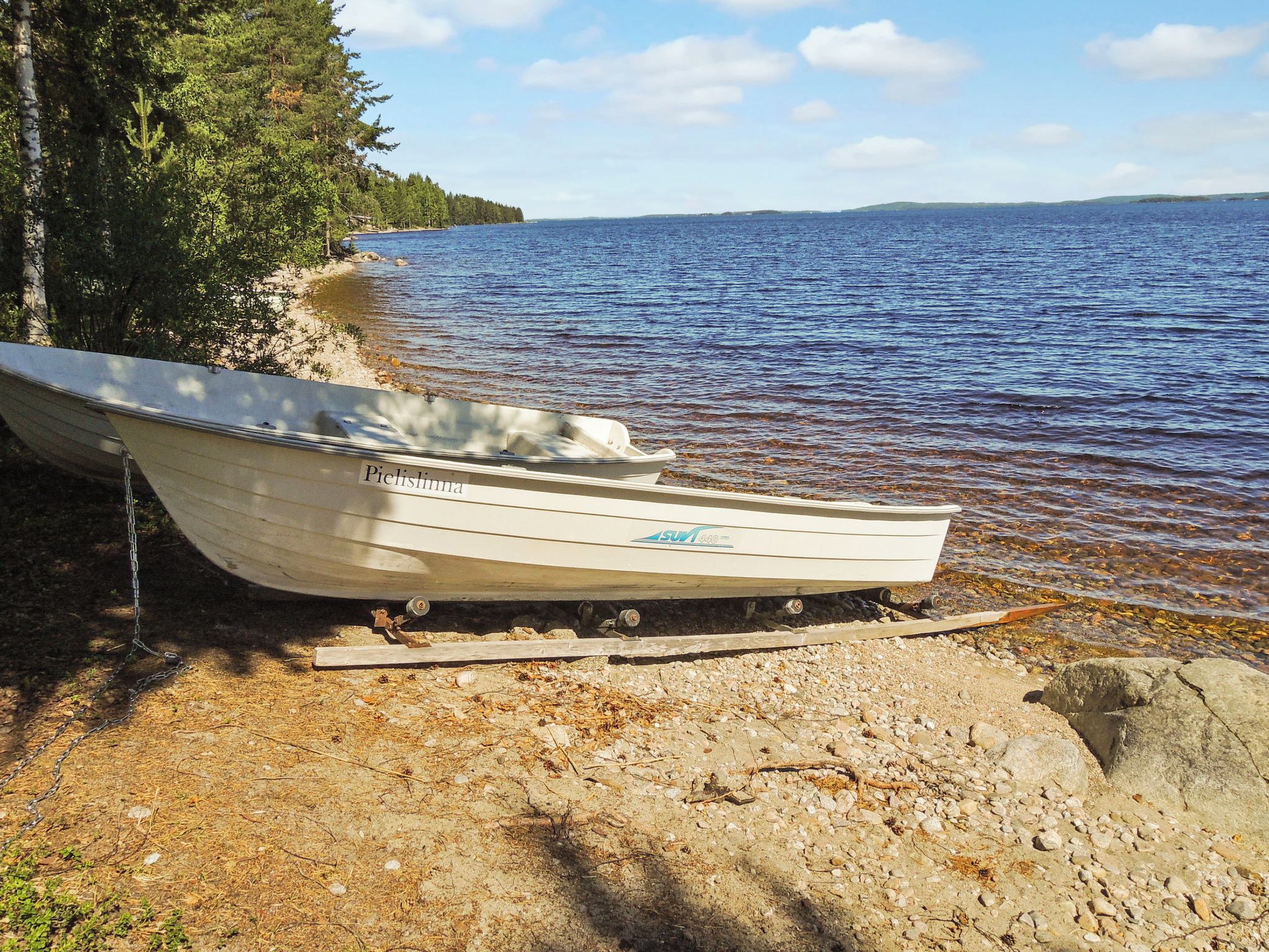 En båt med namnet ”Pielisjoki” låg förtöjd vid en stenig strand, med en skog i bakgrunden och ett stilla vattenområde.
