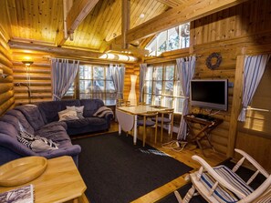 Interior de una cabaña de troncos con un sofá, una mesa de comedor, un televisor y una chimenea.