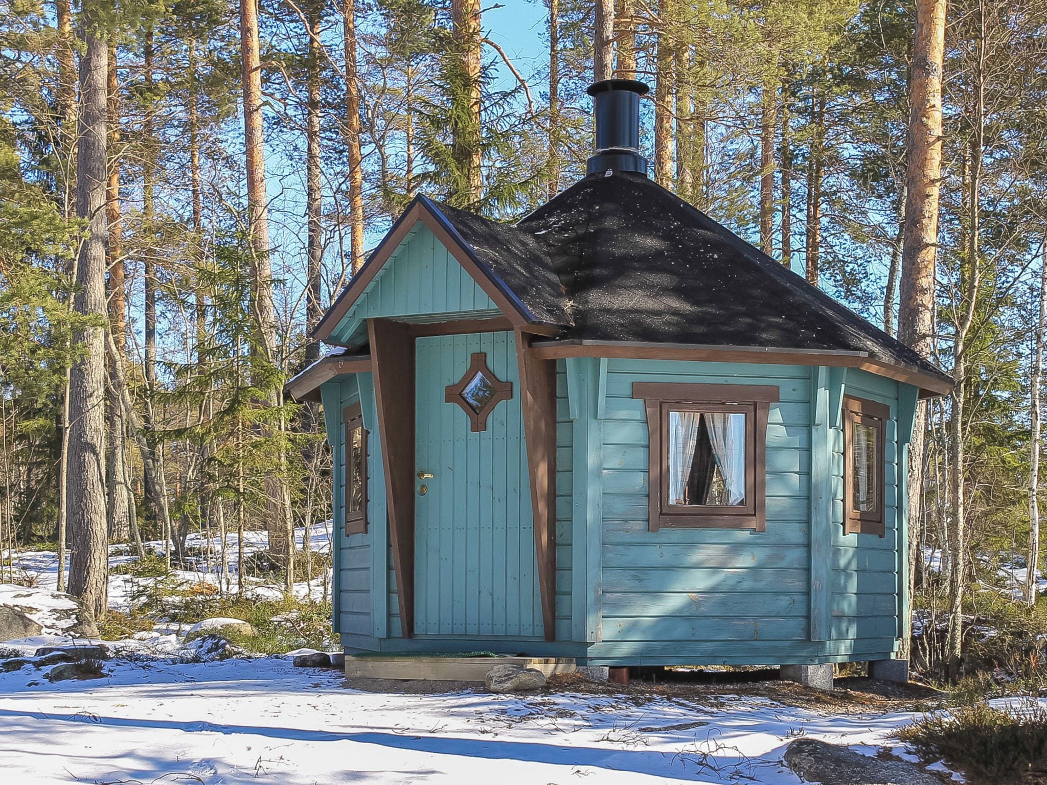 Pieni puinen mökki, jossa on jyrkkä katto, savupiippu ja ikkuna, jossa on verhot.