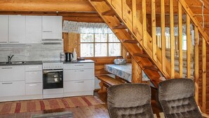 A rustic cabin interior with a wooden staircase, a kitchen with white cabinets, and a dining area with two chairs.