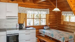 A cabin-style kitchen with a built-in oven, a coffee maker, a kettle, and a radio on the counter. There is a wooden bench and a table with a floral tablecloth.